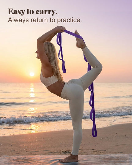 Woman balancing on one leg at the beach, using a purple stretching strap to hold her raised back foot.